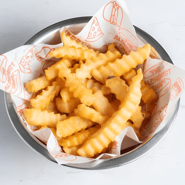 A metal bowl filled with golden, crinkle-cut fries, lined with branded Alchemist Shack paper.