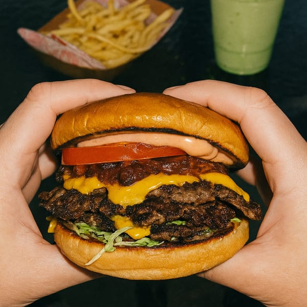Hands holding a double cheeseburger from The Alchemist Shack with fries and a green drink in the background.